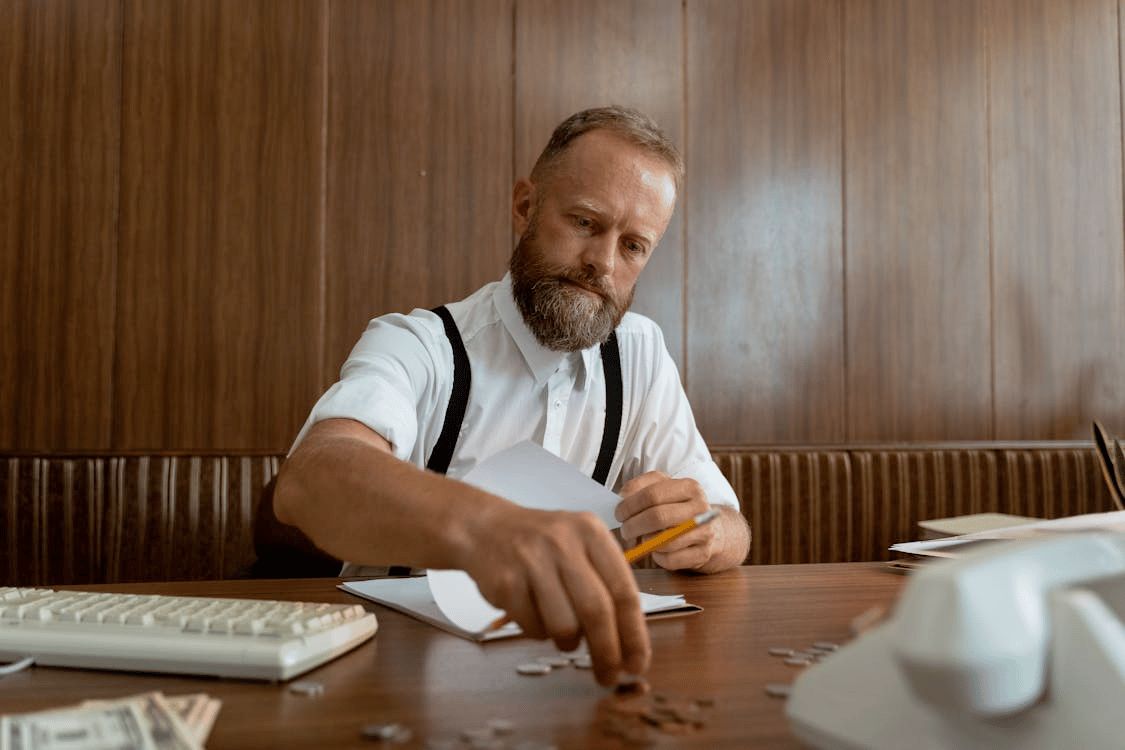 A man at a wooden desk counting coins and notes.